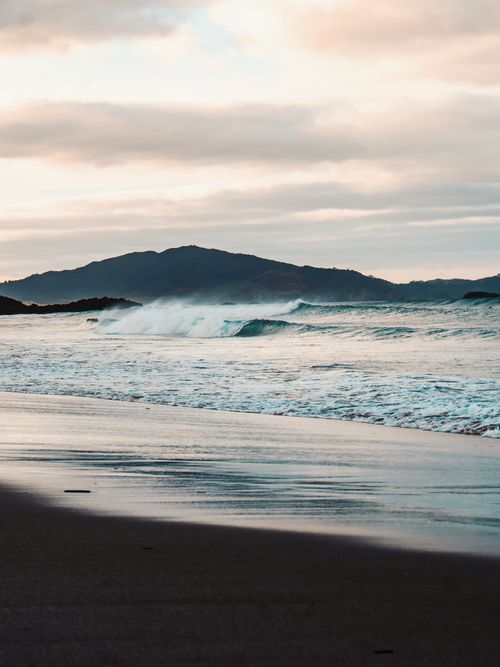 Free Tranquil beach scene in New Zealand with waves and distant mountains at sunset. Stock Photo