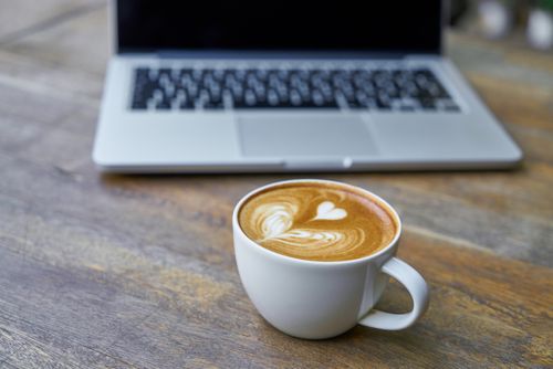 Free A cappuccino with heart-shaped latte art on a wooden table next to a laptop, ideal for work and coffee themes. Stock Photo