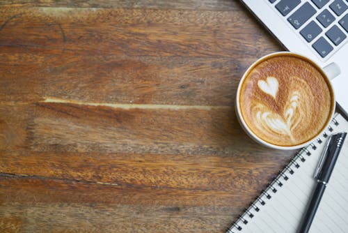 Cup of fresh cappuccino near laptop keyboard and notebook