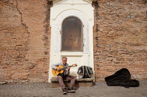 A Man Playing a Classical Guitar While Sitting on the Street Near the Arch Window