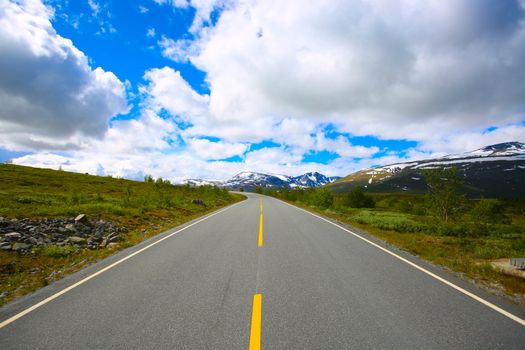 Empty asphalt roadway running along green terrain and leading to mountains covered with snow under bright sky