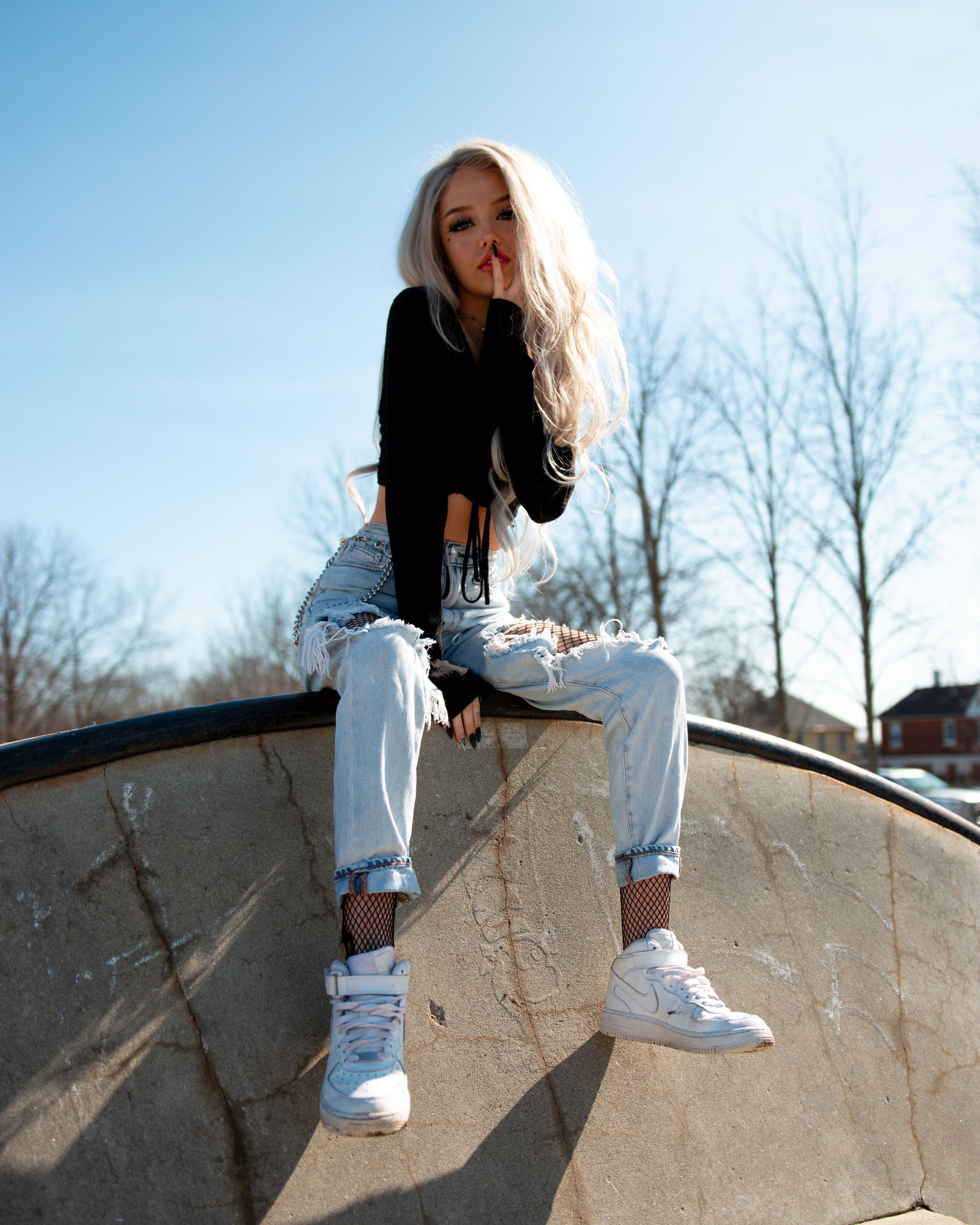 Blonde woman in casual attire sitting on a wall under a blue sky in a modern outdoor setting.