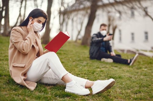 Free Side view of concentrated female in medical mask and coat sitting on lawn in park and reading book on blurred background background Stock Photo