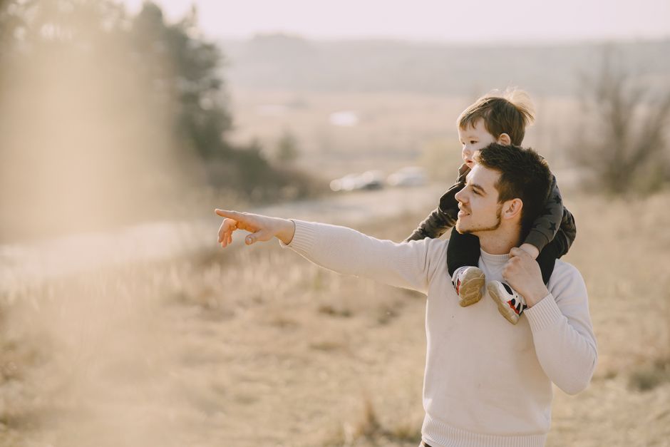 Un padre lleva a su hijo sobre los hombros, señalando hacia el horizonte en un día soleado al aire libre.