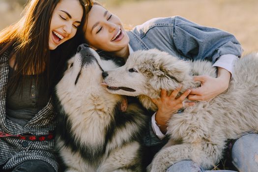 Cheerful female friends with closed eyes in casual wear embracing gently funny fluffy dogs while laughing and sitting in back lit on sunny day