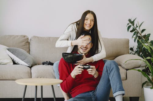 Happy girlfriend covering unrecognizable mans eyes with hand while using game pad and sitting on cozy sofa with bowl at home near potted plant