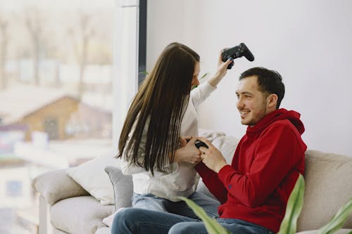 Unrecognizable woman playing with cheerful bearded boyfriend using game pad while sitting on sofa and having fun in apartment in daylight