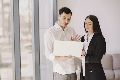 Young male employee with laptop talking to well dressed female coworker while standing near sofa and window in comfortable office