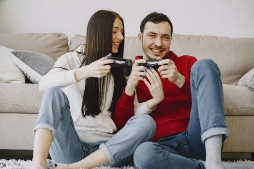 Cheerful man and girlfriend in jeans playing game console while sitting on soft rug leaned on sofa spending time together in apartment