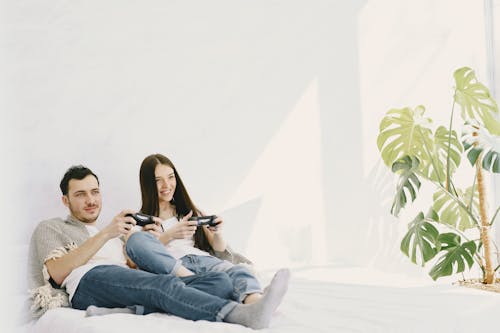 Content man in jeans with game pad lying near positive girlfriend on bed near potted plant on sunny day while entertaining together