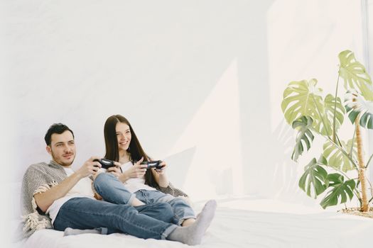 Content man in jeans with game pad lying near positive girlfriend on bed near potted plant on sunny day while entertaining together