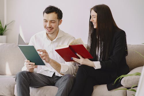 Cheerful male and female coworkers in formal clothes sitting on couch and holding notebooks in hands while laughing and working