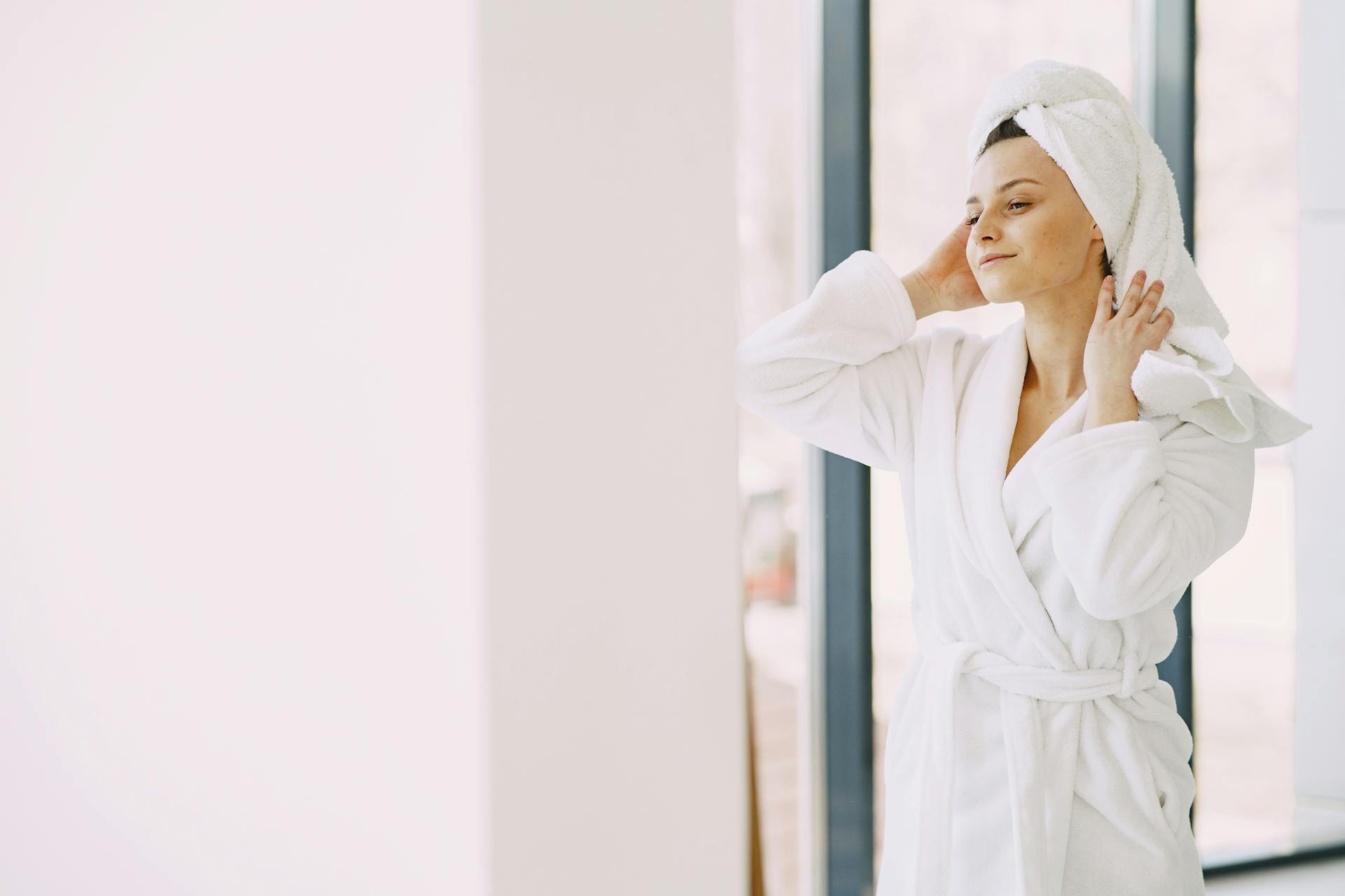 Pensive female in white bathrobe and towel on head smiling and standing near glass window in living room while looking away