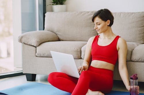Free Smiling female with short hair in activewear sitting near sofa with bottle of water on mat in living room and browsing laptop Stock Photo
