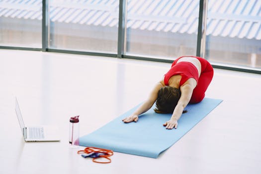 Flexible sportswoman in red sportswear practicing child pose on blue sport mat during workout in modern sport club or gym with glass wall adhering to healthy lifestyle while using netbook for listening meditative music