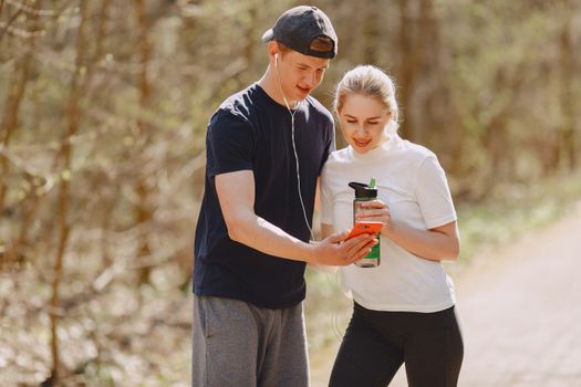 Young man in earbuds sharing cellphone with young woman drinking water from plastic bottle during workout in nature together