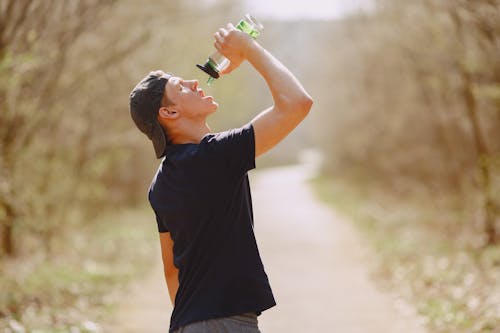 Side view of young male athlete wearing sports clothes and cap drinking water from plastic bottle on blurred background of forest during running and workout