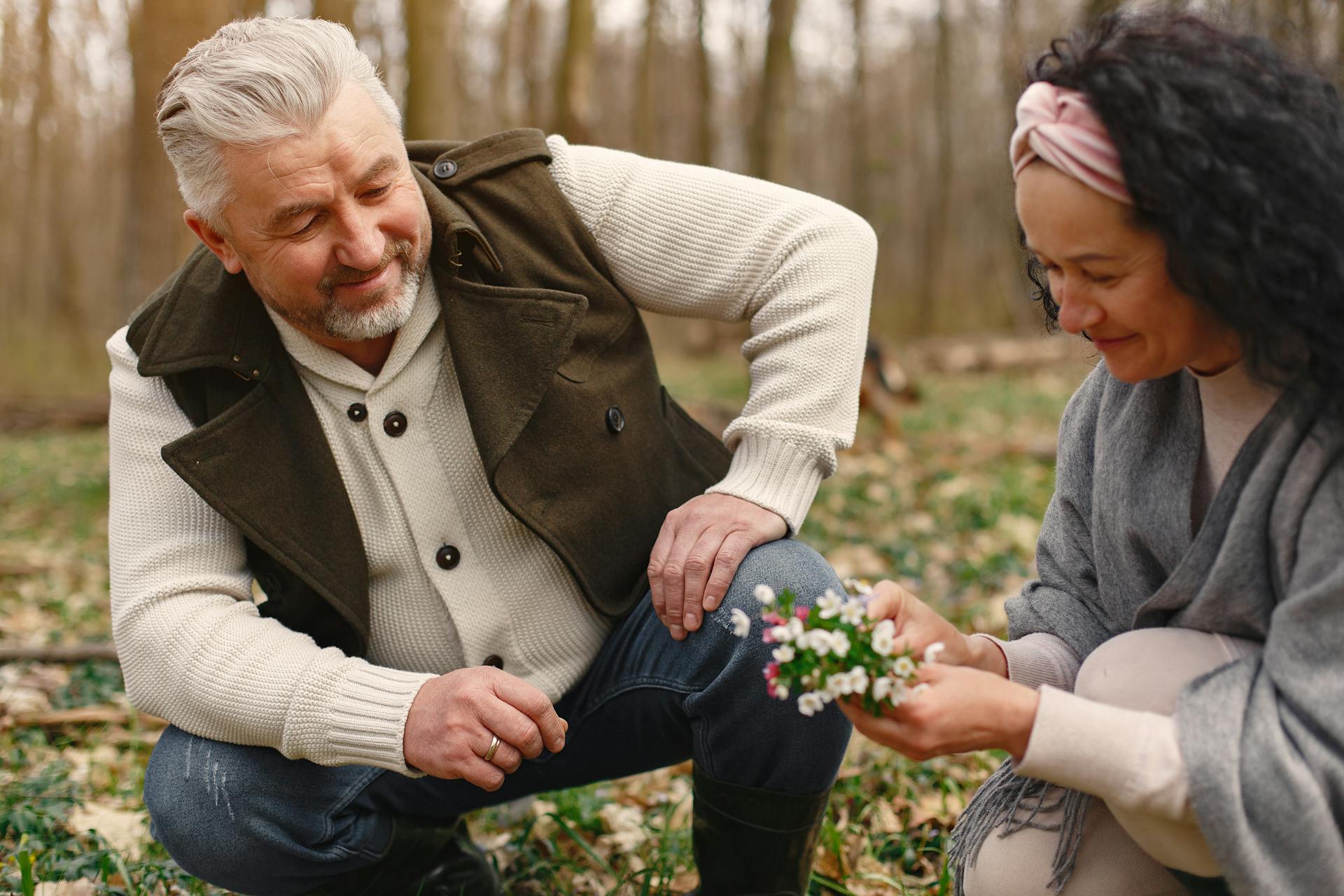 Happy senior wife wearing wide scarf and headband smiling while sitting on ground in autumnal forest with delicate flowers in hands near cheerful elderly gray haired husband in warm clothes while strolling together