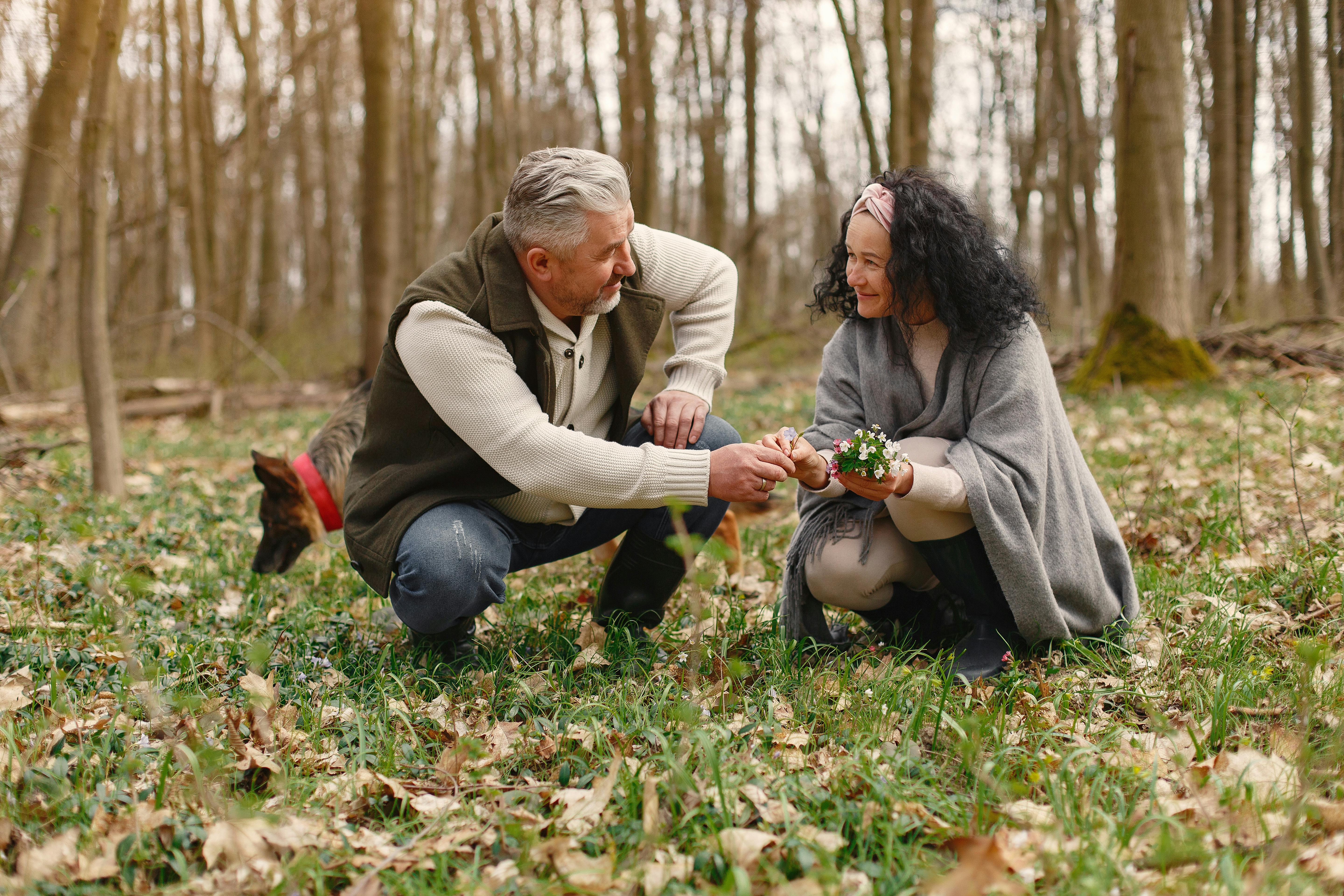 Senior wife wearing wide scarf and headband smiling while sitting on ground in autumnal forest with delicate flowers in hands near elderly gray haired husband in warm clothes while walking with German Shepherd dog and looking at each other