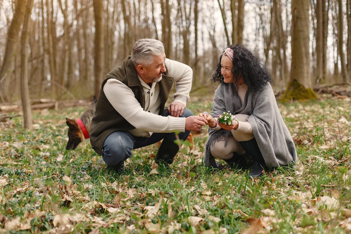 Senior wife wearing wide scarf and headband smiling while sitting on ground in autumnal forest with delicate flowers in hands near elderly gray haired husband in warm clothes while walking with German Shepherd dog and looking at each other