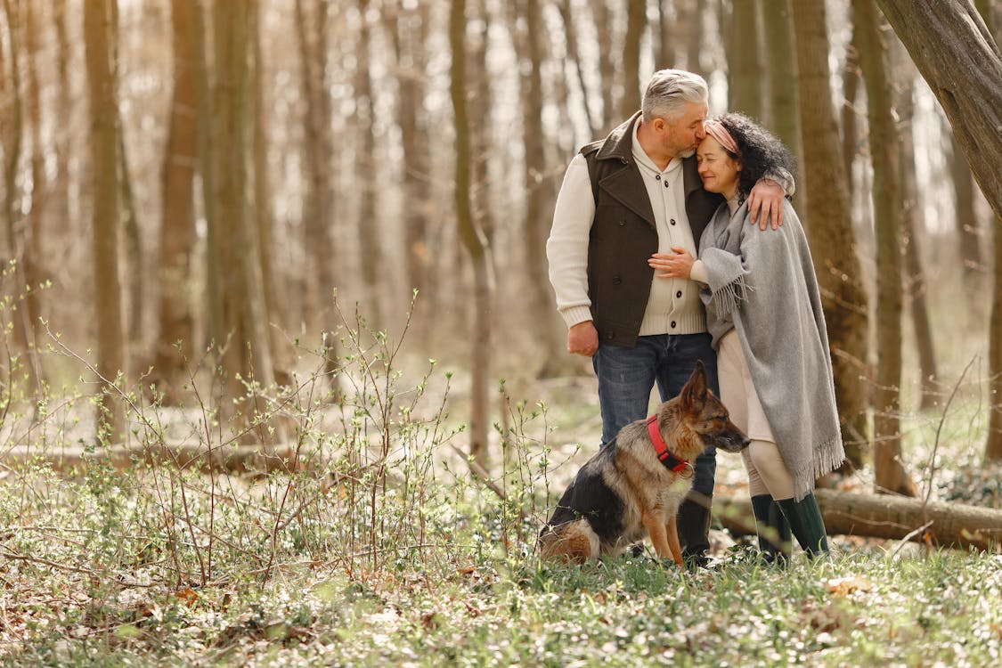Elderly Couple Standing in The Woods On A Bright Day With Their German Sheperd