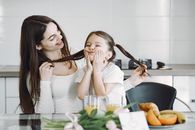 Cheerful mother and daughter in domestic clothes sitting together at table and playing with each other while enjoying breakfast in modern kitchen