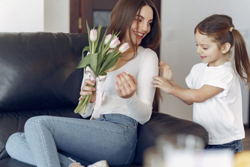Free Delighted young woman in trendy wear smiling and taking bouquet of tulips from daughter in casual clothes while resting in cozy living room during mother day Stock Photo