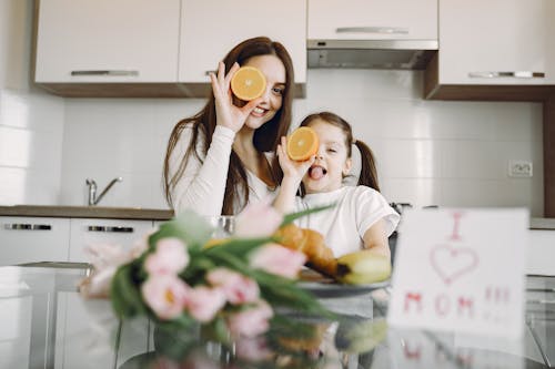 Free From below of cheerful mother and daughter in domestic clothes smiling and playing with oranges while sitting together at table with bouquet of tulip and drawing of child Stock Photo