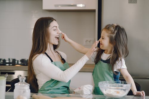 Side view of cheerful young mother and little daughter in aprons cooking pastry and laughing while smearing noses each other with dough on light modern kitchen