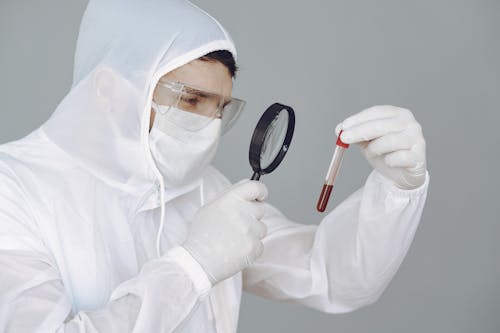Person Wearing Personal Protective Equipment While Holding Magnifying Glass and Test Tube