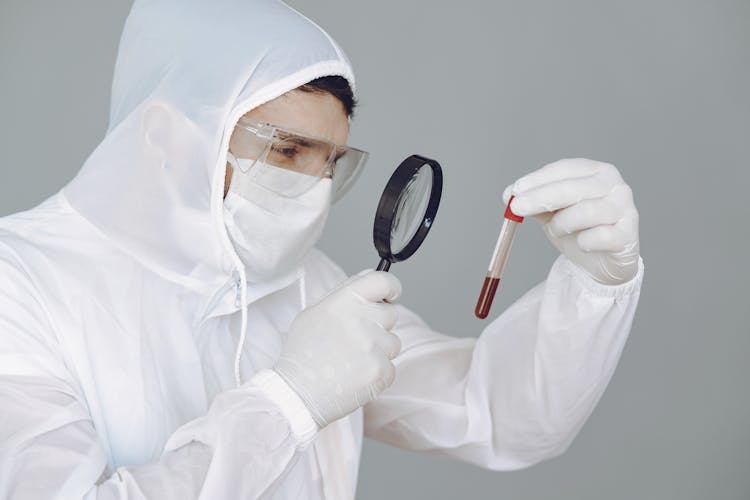 Person Wearing Personal Protective Equipment While Holding Magnifying Glass And Test Tube