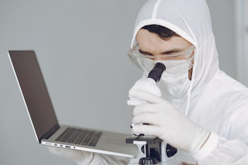 Person in Personal Protective Equipment Holding a Laptop While Using Microscope