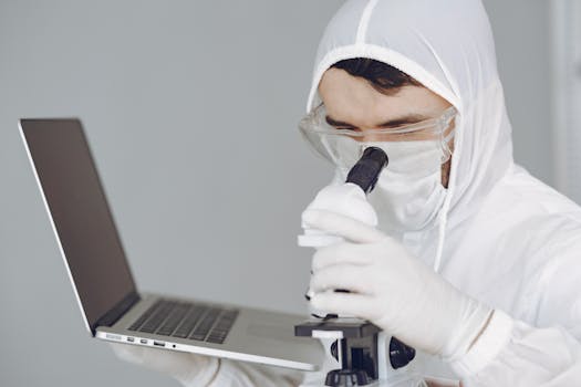 Scientist in protective gear analyzing samples with a microscope and laptop.