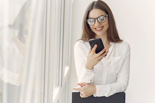 Cheerful young female in trendy clothes and eyeglasses messaging on mobile phone while standing near window and leaning on chair in modern light office