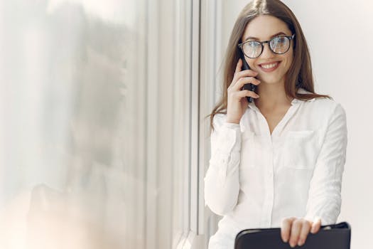 Cheerful woman in glasses talking on smartphone by a window indoors.