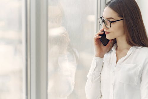 Free Pensive young woman in casual wear and eyeglasses speaking on mobile phone and looking at window while resting in modern apartment Stock Photo