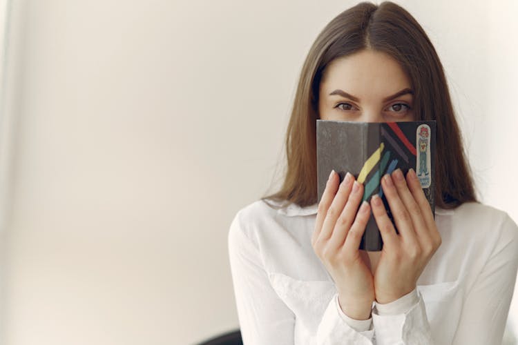 Charming Woman Covering Face With Notebook In Office