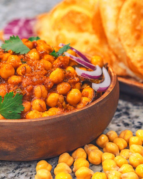 Appetizing tasty stewed chickpeas in bowl topped with red onion slices and parsley placed on marble table near plate with fresh bread