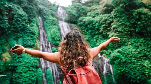 From below back view unrecognizable female tourist with backpack standing with arms outstretched and enjoying wonderful scenery of waterfall in green tropical rainforest