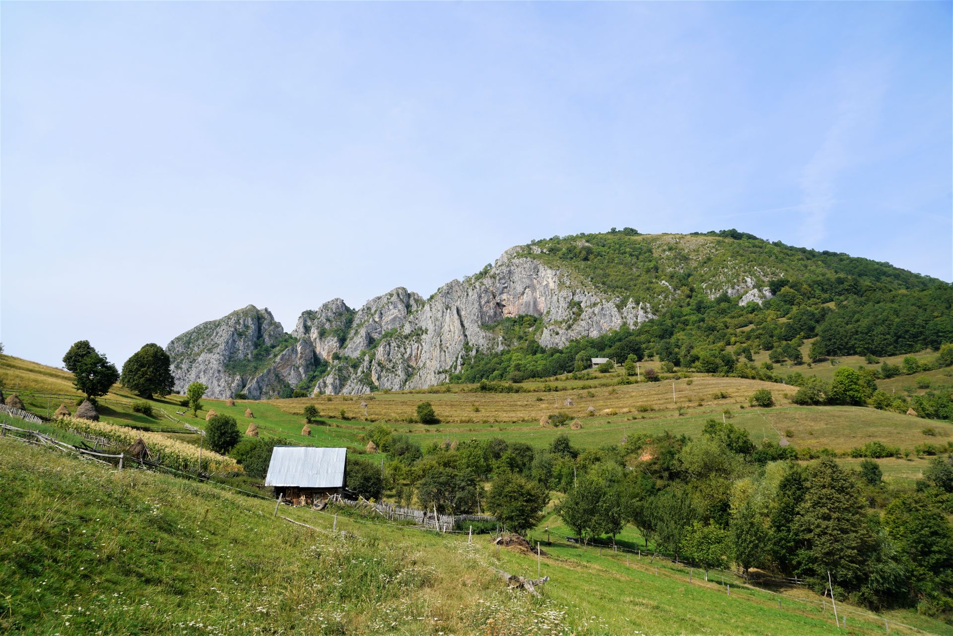 Scenery view of small countryside house roof on hillside with grass and growing trees behind rough mounts under blue cloudy sky in summer