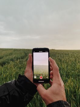 Hands holding a smartphone to photograph a vast green field in a rural setting during the day.