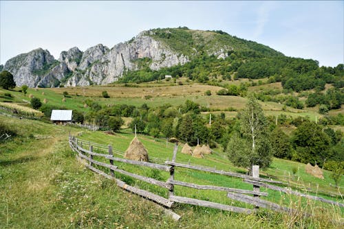 Picturesque view of hill with grass and wooden fence with haystacks behind small rural house and mountains under blue cloudy sky