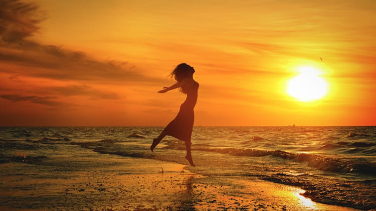 Silhouette Of Unrecognizable Woman Jumping Above Sea Beach At Sunset