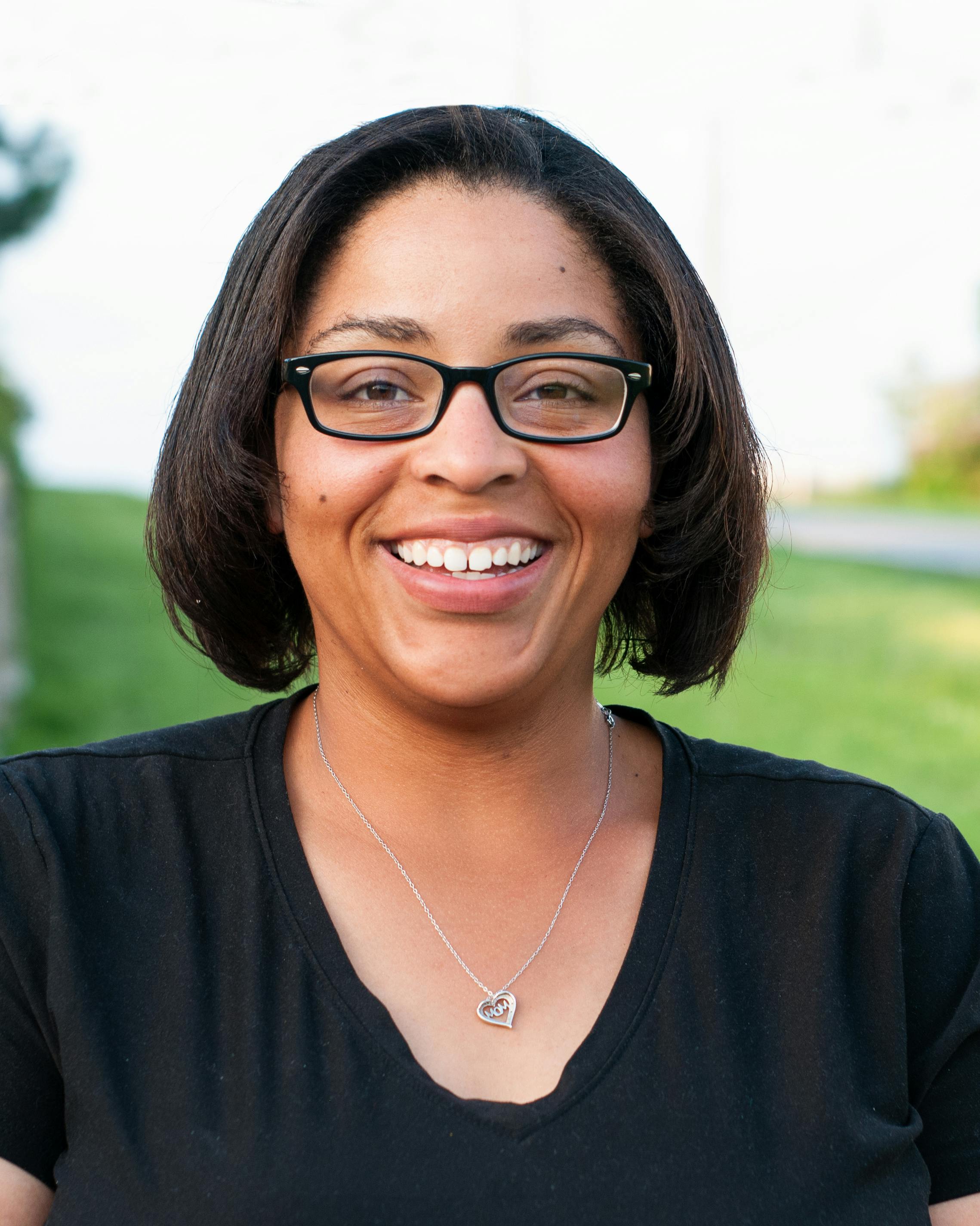 Positive middle aged ethnic female in eyewear and pendant looking at camera while standing on grass meadow under serene sky in daylight in countryside