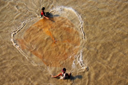 Free Drone view of anonymous ethnic female catching fish with net in brown lake with rippled water in daylight Stock Photo