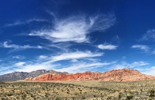 From below of colorful blue sky with clouds under rough mountains and plants growing on hill with dry terrain in daylight