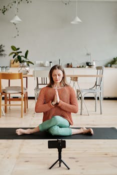 Woman practicing yoga on a mat indoors, with smartphone setup for video.