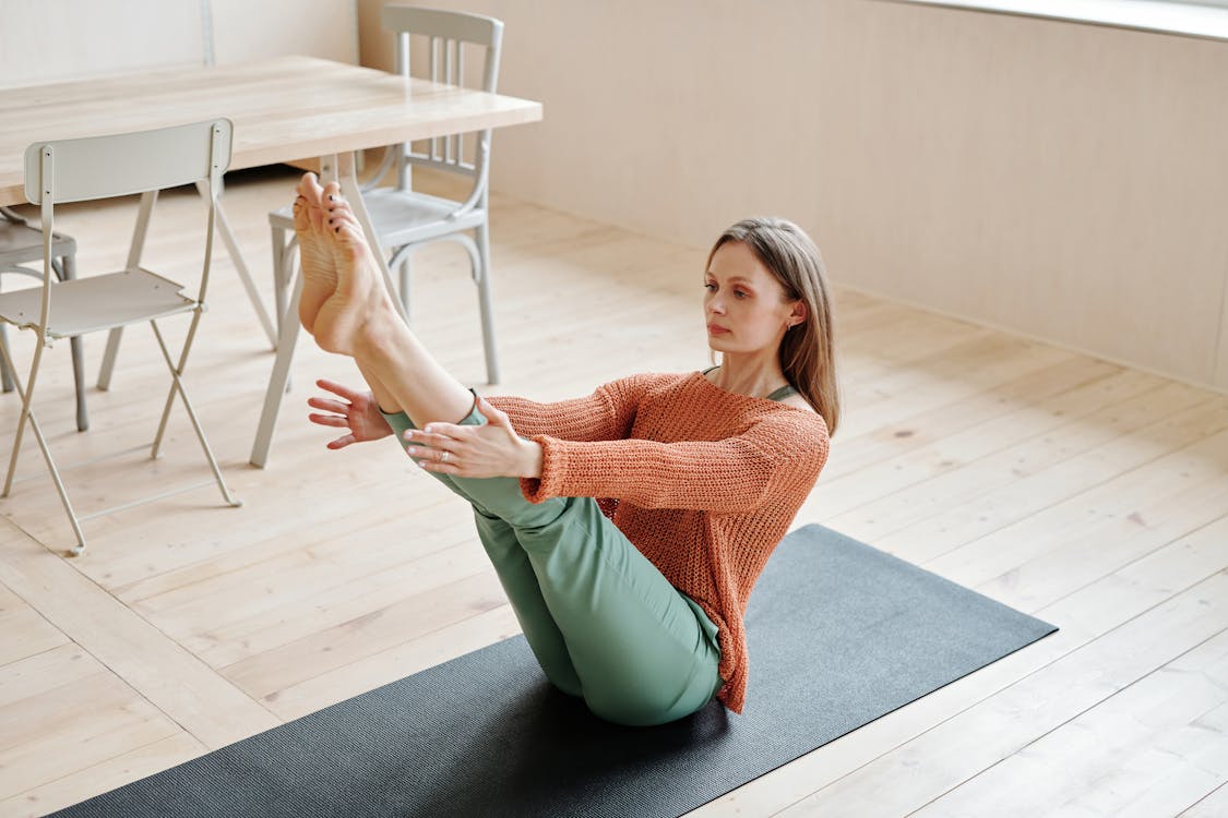 Woman in Orange Knit Sweater and Green Pants Sitting on a Yoga Mat