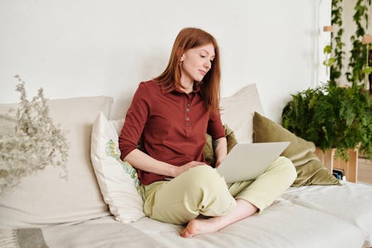 A young woman sits on a couch with a laptop, working remotely in a cozy home setting.
