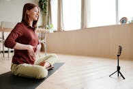 一位年轻女子在家中练习瑜伽，并用智能手机录制课程，自然光从窗户透入